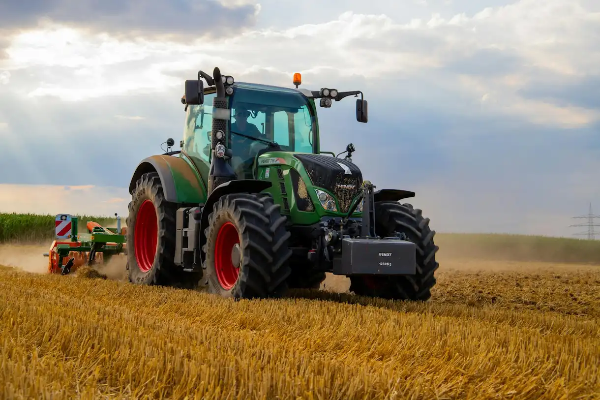Tractor agrícola verde trabajando en un campo de cultivo, con apero enganchado y polvo levantándose al avanzar.